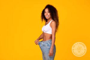 Happy black young lady showing too big jeans and results of successful weight loss diet over yellow background.